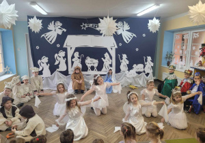 Gwiazdki i aniołki tańczą przy piosence "Przy kominie świerszczyk spał". Dzieci przykucnęły na podłodze z rozłożonymi rękami, w których trzymają białe chustki. W tle dekoracja oraz pozostali uczestnicy przedstawienia.