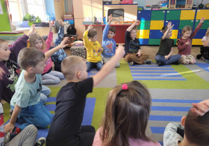 Dzieci siedzą w kółku na dywanie. Przedszkolaki trzymają rączki w górze zgłaszając się do odpowiedzi na zadane pytanie.