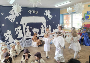 Ala, Julka, Lena, Wiktoria, Łucja i Winek w białych strojach gwiazdek i aniołków tańczą w kółku trzymając się za ręce. Po lewej stronie widać chłopców w strojach pastuszków i baranków.