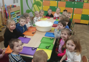Dzieci siedzą przy wspólnym stole, na którym znajduje się miska ze śniegiem, kolorowe tacki i lupy.