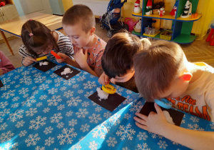 Czworo dzieci siedzi przy stole i obserwuje przez lupy płatki śniegu znajdujące się na czarnych kartonach.