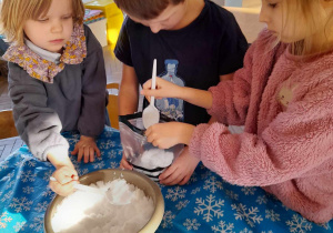 Troje dzieci stoi przy stole, na którym znajduje się miska ze śniegiem. Tomek trzyma foliowy woreczek, a Łucja i Zosia wkładają do niego śnieg za pomocą łyżek.