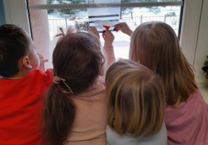 Czworo dzieci stoi przy oknie i dotyka foliowego woreczka, w którym znajduje się woda i guziczki po roztopionym baławanku.