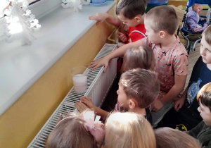 Dzieci przykucnęły przy grzejniku i obserwują jak śnieg w kubku rozpuszcza się pod wpływem ciepła.