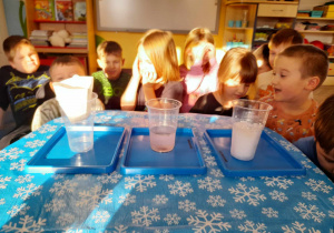 Dzieci siedzą na dywanie przy stoliku, na którym stoją trzy plastikowe kubki. W pierwszym od prawej strony znajduje się śnieg, w drugim powstała ze śniegu woda, na trzecim umieszczony jest papierowy filtr.