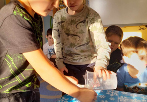 Dwóch chłopców stoi przy stoliku. Eryk trzyma kubek z filtrem, a Samuel przelewa wodę powstałą ze śniegu przez papierowy filtr.