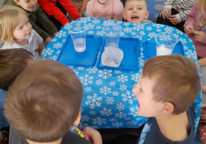 Dzieci siedzą na dywanie wokół stolika i oglądają osad, który pozostał na papierowym filtrze po przelaniu wody powstałej z rozpuszczonego śniegu.