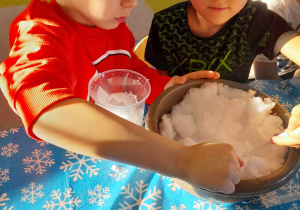Antoś i Eryk siedzą przy stole i wkładają z miski śnieg do plastikowego kubka za pomocą łyżki.