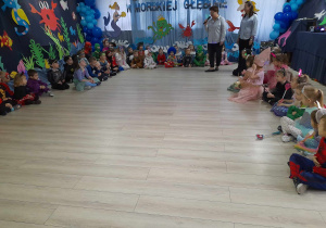 Dzieci w strojach karnawałowych siedzą na podłodze na tle dekoracji pod hasłem "W Morskiej Głębinie", a Pani Karolina i Pani Angelika witają przedszkolaków na balu.