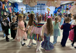 Dzieci w strojach karnawałowych tańczą we wspólnym kole. W tle dekoracja balowa.