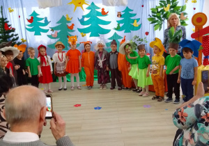 Grupa "Biedronek" w półkolu w strojach postaci do Czerwonego Kapturka podczas występu z okazji Dnia Babci i Dziadka. W tle dekoracja: na niebieskim tle drzewa, ptaki, owady i słońce.