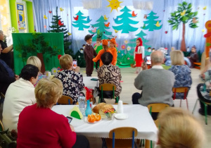 Grupa dzieci na tle dekoracji w przebraniach: jeża, wiewiórek i Czerwonego Kapturka podczas występu. Przy stolikach babcie i dziadkowie oglądają popisy swoich wnucząt.