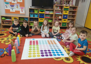 Dzieci siedzą w półkolu na dywanie, przed nimi leży mata do kodowania i kolorowe kubeczki.