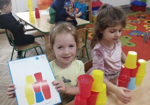 Emma prezentuje ustawioną wieżę z kubków w kolorach niebieskim, czerwonym, żółtym oraz kartę wzoru. Obok koleżanka buduje swoją wieżę.
