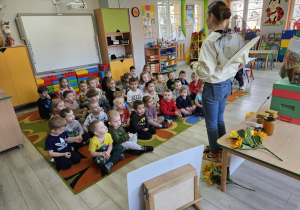 Pani Justyna opowiada dzieciom o pszczołach stoją przed nimi. Dzieci siedzą na dywanie.