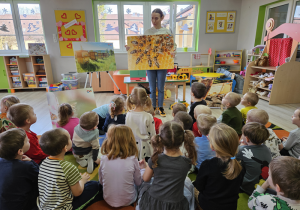 Pani Justyna stoi przed dziećmi siedzącymi na dywanie, w rękach trzyma ilustrację pszczół, o których opowiada.