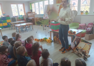 Pani Justyna prezentuje dzieciom ilustracje pszczoły, na stoliku inne akcesoria potrzebne na warsztaty.