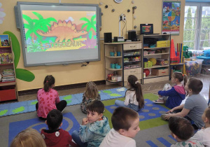 "Słoneczka" siedzą na dywanie i oglądają film edukacyjny na tablicy multimedialnej.