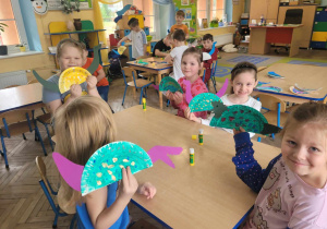 Dzieci z grupy "Słoneczek" siedzą przy stolikach w trakcie wykonywania pracy plastycznej. Przedszkolaki tworzą wizerunki dinozaura z papierowych talerzyków.