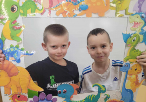 Kacper i Antoś uśmiechają się do zdjęcia przy foto-ramce z wizerunkami dinozaurów.