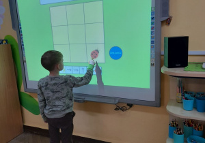 Samuel stoi przed tablicą multimedialną. Chłopiec prowadzi biedronkę na planszy zgodnie z kodem.