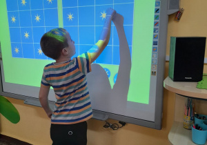 Franek stoi przed tablicą multimedialną. Chłopiec układa gwiazdki na planszy zgodnie z podanym wzorem.