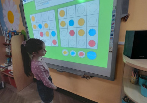 Julka stoi przed tablicą multimedialną podczas zabawy "Kolorowe kółka". Dziewczynka ułożyła na planszy wzór z kolorowych kółek i sprawdza poprawność wykonanego zadania.