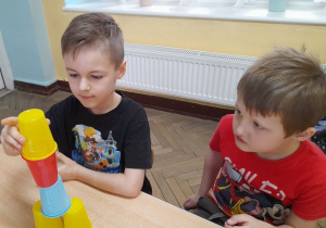 Przy stole siedzi Oskar i Tomek. Oskar dokłada żółty kubek na wieżę.