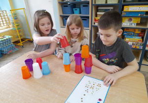 Przy stole siedzą Ala, Lena i Franek. Dzieci budują wieżę z kolorowych kubków. Przed nimi leży karta z zakodowanym wzorem.