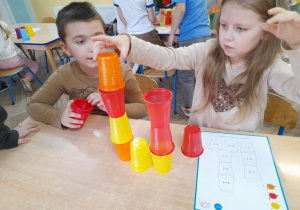 Gabryś i Gabrysia siedzą przy stole. Dzieci wykonują działania przedstawione na karcie wzoru i budują według kodu wieżę. Dziewczynka dostawia pomarańczowy kubek.