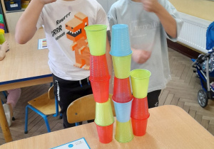 Kacper i Antoś stoją przy stole. Chłopcy prezentują swoją wieżę oraz kartę z zakodowanym wzorem.