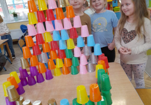Gabryś, Anastazja i Gabrysia stoją obok stołu i prezentują wieżę zbudowaną z kolorowych kubków według własnego pomysłu.