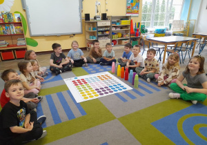Dzieci siedzą na dywanie w półkolu, a na środku leży mata do kodowania. Obok maty stoją kolorowe kubki.