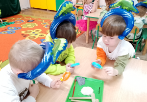 Grupka dzieci w opaskach z dinozaurami siedzi przy stoliku i obserwuje przy użyciu lup wykopaliska.