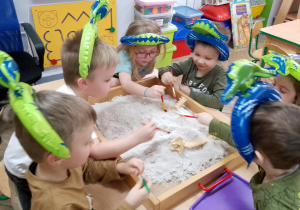 Grupka dzieci w opaskach z dinozaurami siedzi przy stoliku i wykopuje z piasku szkielet dinozaura.