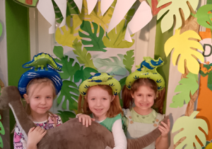 Nikola, Emma i Nadia w opaskach dinozaurów trzymają dinozaura i pozują do zdjęcia na foto ściance w paszczy zielonego dinozaura. W tle zielone liście.