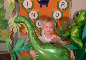 Wiktor na tle dekoracji pozuje do zdjęcia trzymając zielonego dinozaura.