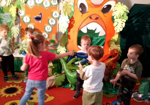 Wiktor i Mikołaj stoją z tacami na których znajdują się opaski z dinozaurami i rozdają je dzieciom. W tle dekoracja z okazji Dnia dinozaura.