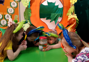 Grupka dzieci przy zielonym stoliku z kolorowymi lupami obserwuje wykluwającego się dinozaura w słoiku wypełnionym wodą.