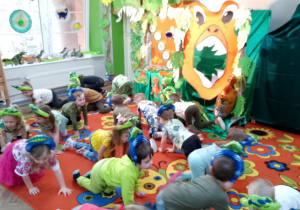 Dzieci w opaskach z dinozaurami chodzą na czworakach naśladując dinozaury. W tle dekoracja z okazji Dnia dinozaura.