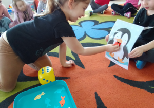 Lena karmi rybkami pingwina. Obok leży kostka do gry, która wskazuje ilość rybek, które ma zjeść pingwin.