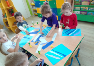 Dzieci wycinają paski papieru w różnych odcieniach niebieskiego i naklejają na karton, co będzie tłem dla pingwina.