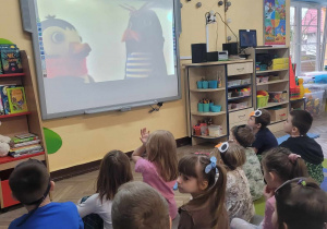 Dzieci z grupy Słoneczek siedzą na dywanie i oglądają film edukacyjny na tablicy multimedialnej