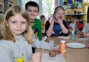 Przedszkolaki z grupy Słoneczek siedzą w sali przy stolikach i jedzą zakupione łakocie: pyszne wypieki i jogurty