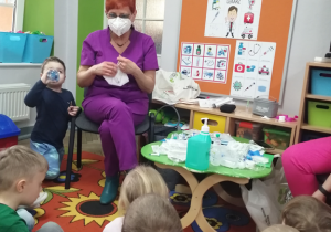 Pani pielęgniarka w maseczce ochronnej tłumaczy dzieciom do czego ona służy.