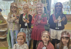 Dziewczynki z grupy w koronach na głowie pozują do zdjęcia trzymając w dłoniach upominki. W tle widać dekorację w sali.