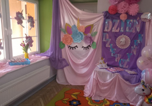 Dekoracja z okazji "Dnia Dziewczynek": na tablicy pokrytej fioletowym materiałem przypięty jest napis "Dzień Dziewczynek" oraz obrazek jednorożca, nad tablicą wiszą różowe bibułowe kwiaty. Przed tablicą na szafce okrytej różowym materiałem na tacach leżą upominki. Z boku stoją balony z nadrukiem jednorożca. Z drugiej strony na różowym materiale przypięty jest obrazek jednorożca. Na oknie wiszą balony w kształcie jednorożca oraz stoją fioletowe tulipany.