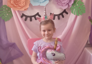 Weronika siedzi na różowym krzesełku i trzyma balon w kształcie jednorożca. W tle dekoracja z okazji "Dnia Dziewczynek".