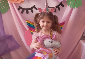 Nadia siedzi na różowym krzesełku i trzyma balon w kształcie jednorożca. W tle dekoracja z okazji "Dnia Dziewczynek".