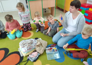 Dzieci razem z Panią Kasią oglądają na dywanie przybory krawieckie: nici, nożyczki, igły, suwaki i inne, obok leża tkaniny.